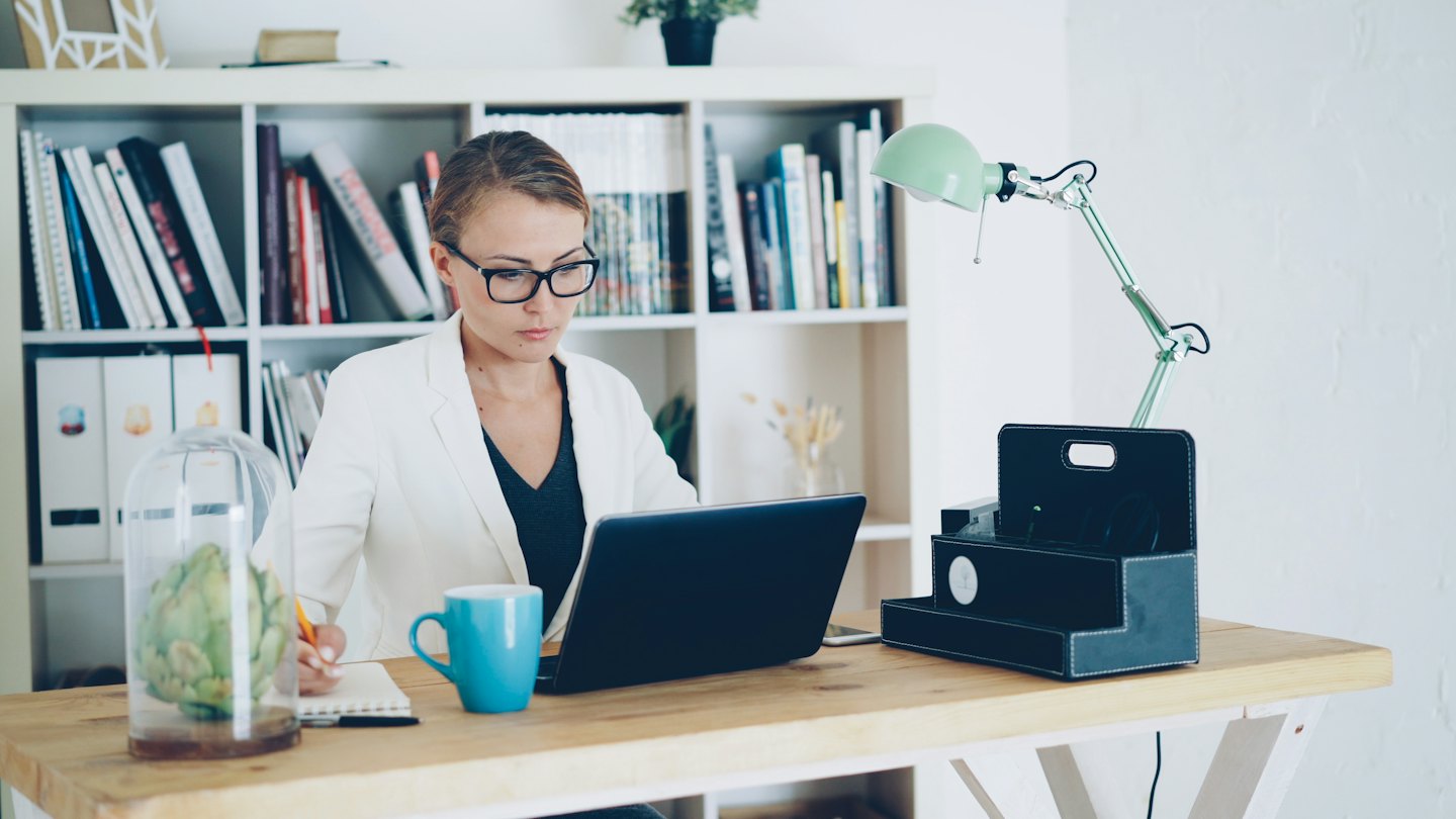 Woman in white blazer types on her laptop at a desk.